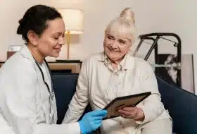 A caregiver shows an older woman a photo on a tablet while they sit together on a sofa.