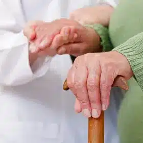 A person wearing a green sweater holds a wooden cane, while another person in a white coat gently supports their hand.