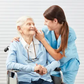A smiling caregiver in blue scrubs gently touches the face of an elderly woman seated in a wheelchair.