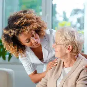 A caregiver with curly hair smiles warmly at an elderly woman wearing glasses and a cardigan.