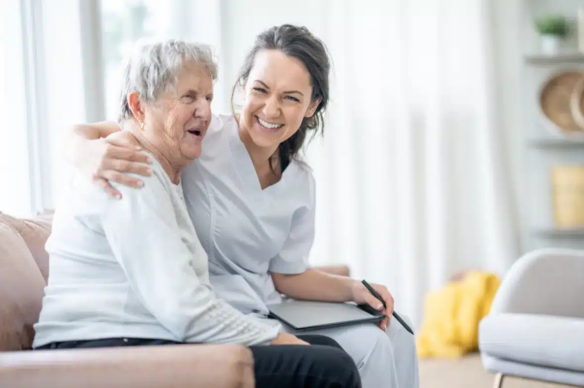 A caregiver in white scrubs warmly embraces an elderly woman on a couch, both smiling joyfully. The atmosphere is bright and comforting.
