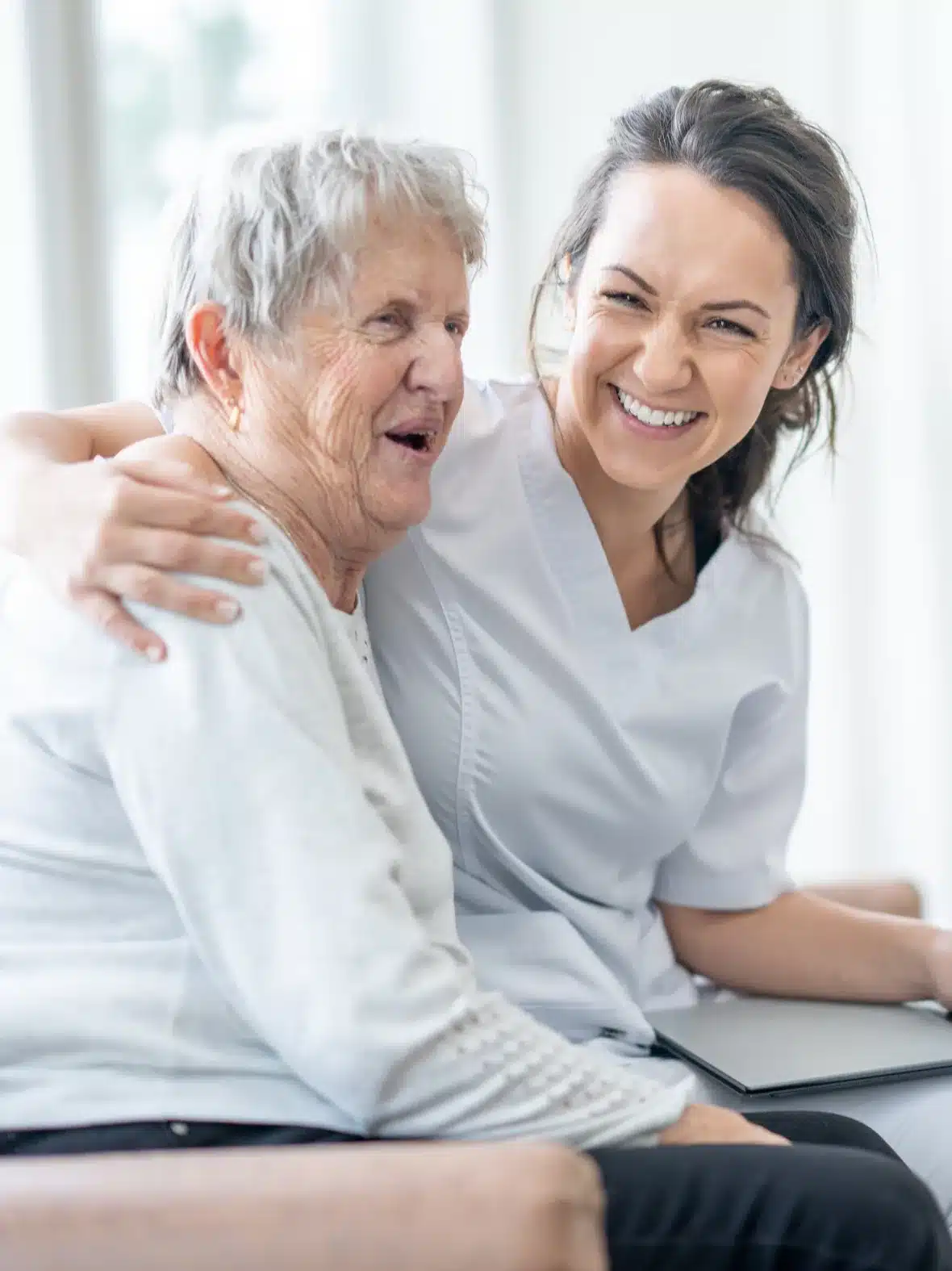 A smiling caregiver hugs an older woman in a bright living room.