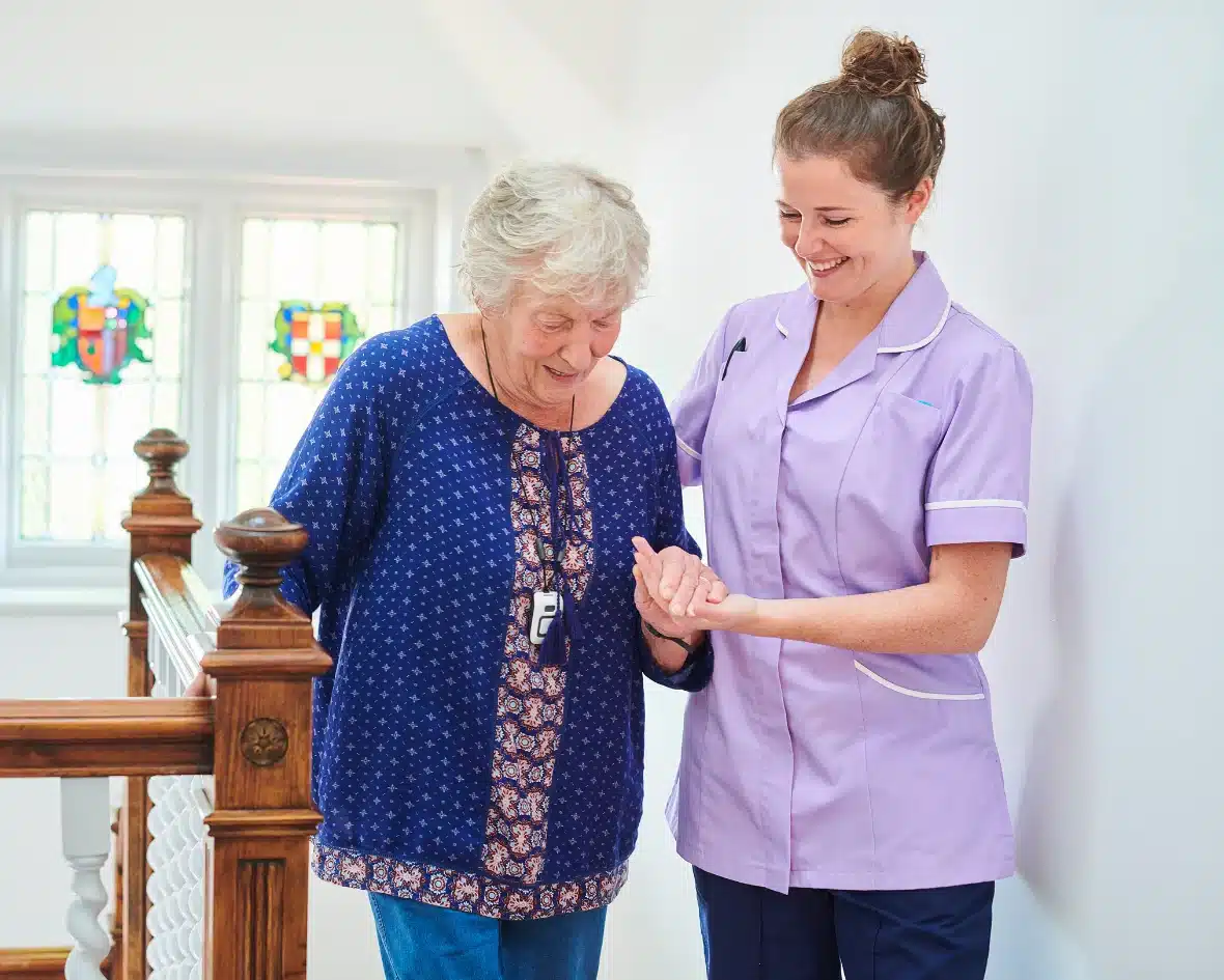 A caregiver in a purple uniform helps an elderly woman down a staircase, both smiling.